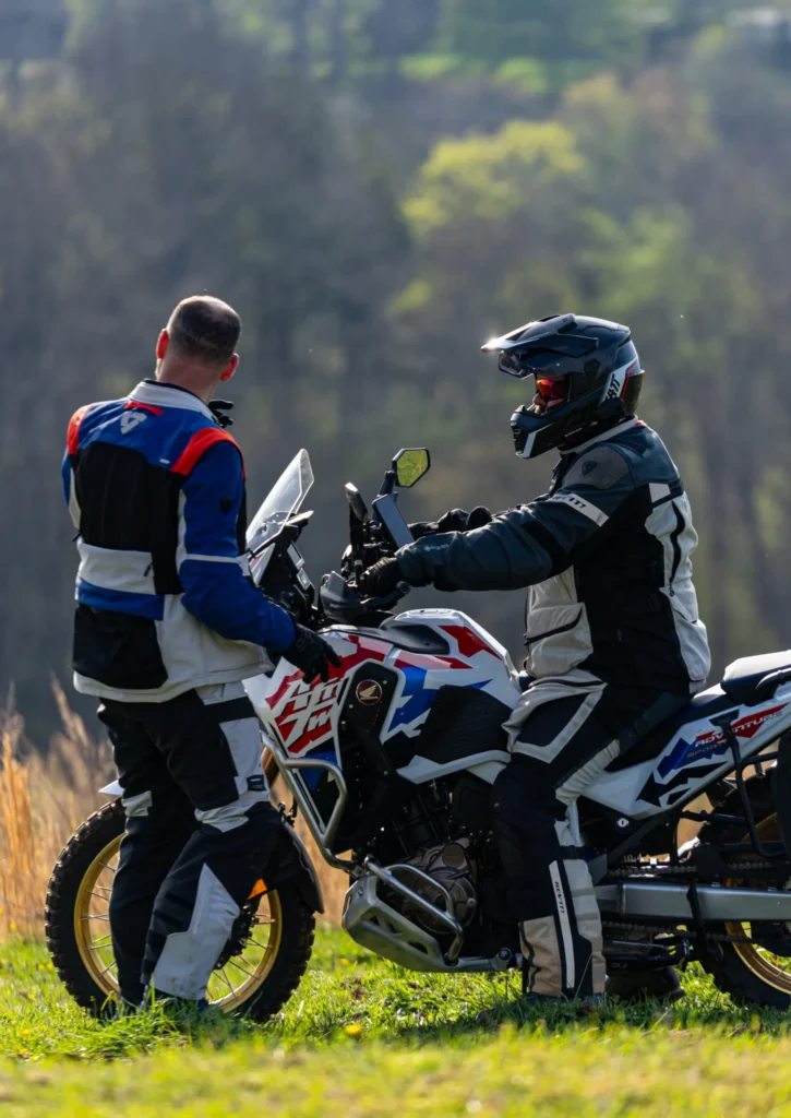 Two motorcyclists in protective gear stand beside dirt bikes in a grassy field.